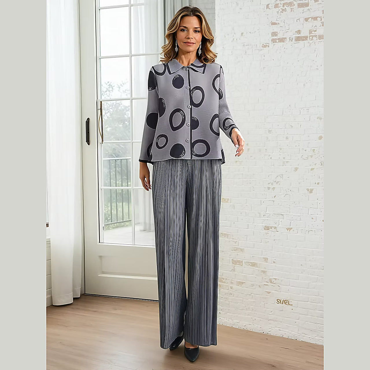 Woman wearing a gray patterned shirt and pants standing in a room with a white brick wall.